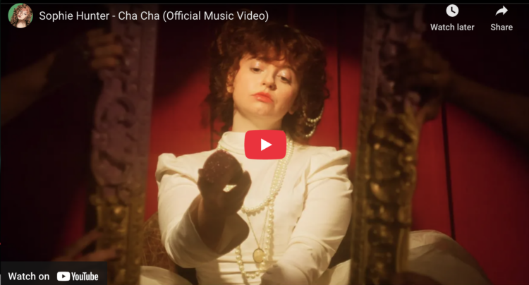 Screenshot of Sophie Turner's video for her song "Cha Cha" showing the artist in a Victorian-inspired dress looking at herself in a mirror compact